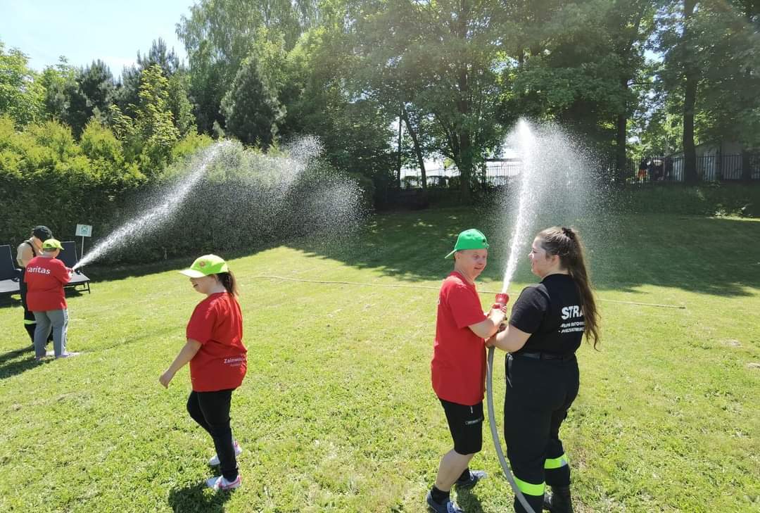 Na zdjęciu widać osoby i strażaków lejących wodę z węża w ogrodzie.