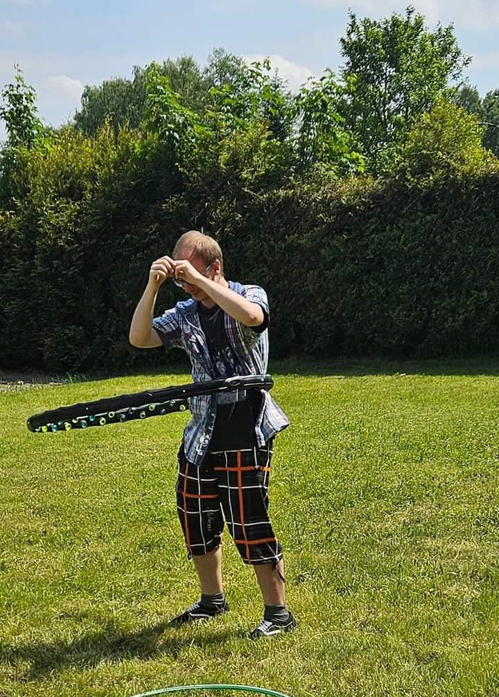 Na zdjęciu widać Uczestnika stojącego na trawie i kręcącego hula hopem w tle ogród.