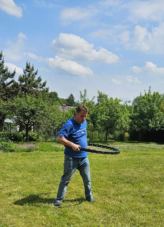 Na zdjęciu widać Uczestnika stojącego na trawie i kręcącego hula hopem w tle ogród.