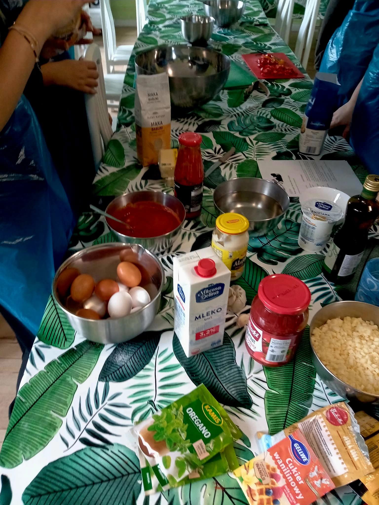 Na zdjęciu widać stół z produktami spożywczymi (mleko, jajka, mąka, przyprawy) , miskami i narzędziami do pracy oraz osoby stojące przy nim.