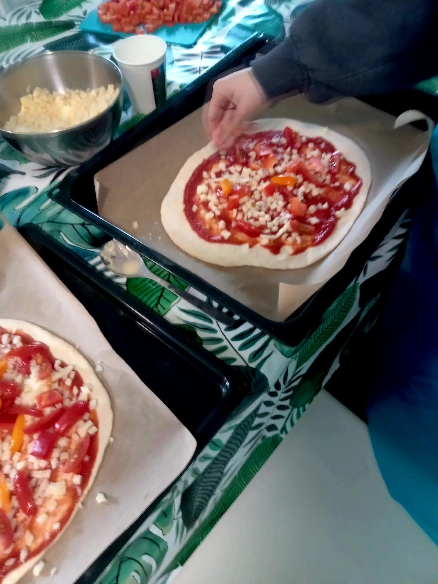 Na zdjęciu widać blachę, na której leży pizza z sosem pomidorowym, serem, papryką itp i rękę posypującą ją kolejnymi dodatkami.