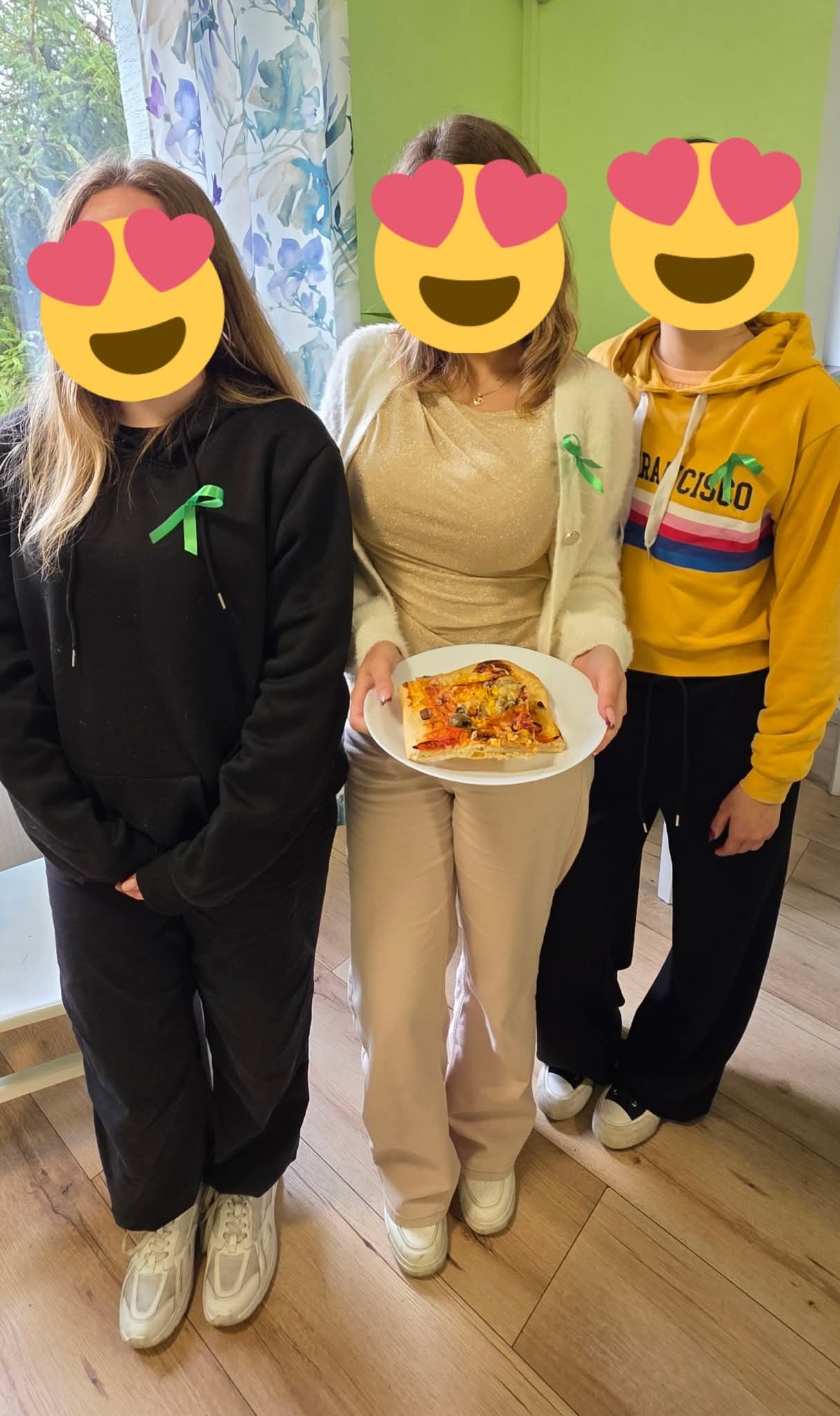 Na zdjęciu widać trzy dziewczyny z zasłoniętymi buźkami twarzami, jedna z nich trzyma talerz z pizzą, mają przyczepione zielone wstążki.