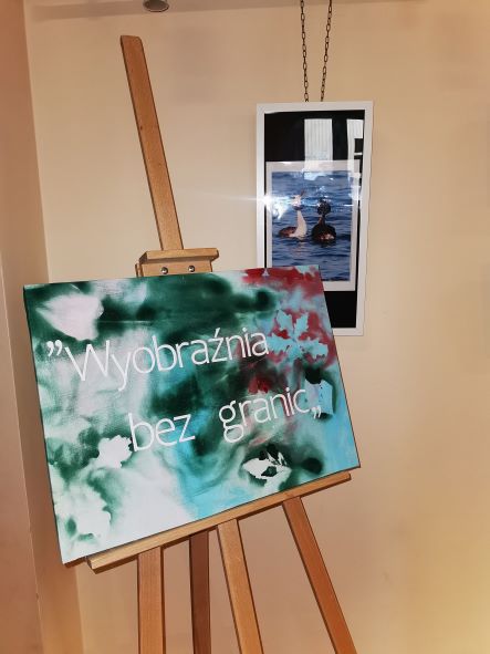 Na pierwszym planie stoi sztaluga z plakatem, na którym widać tytuł wystawy "Wyobraźnia bez granic", w tle zdjęcie przedstawiuające dwa perkozy. 