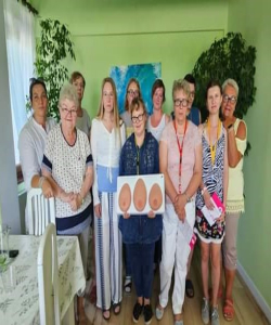 Na zdjęciu widać grupę kobbiet pozujących do zdjęcia. Kobieta stojąca w środku trzyma tablicę z trzema sztucznymi piersiami służącą do nauki badania piersi.