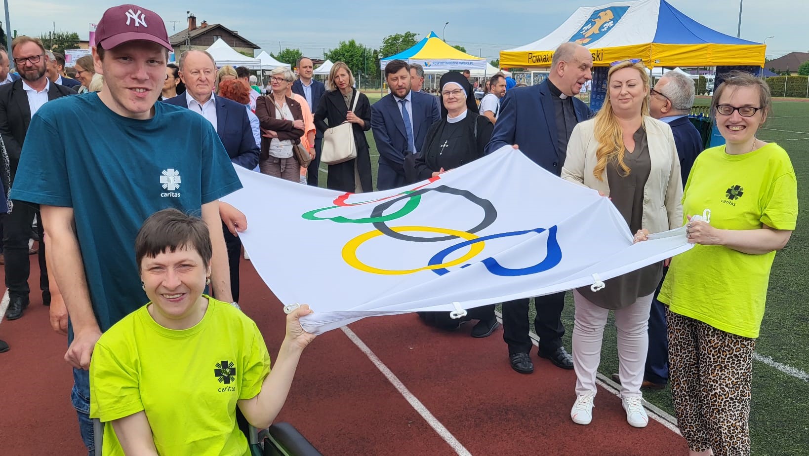 Na zdjęciu widać osoby stojące na bieżni trzymające flagę olimpijską. W tle stadion i biało-żółto-niebieskie namioty.